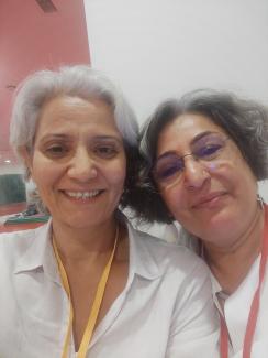 Sultan Çamur Karataş ve Nursel Denir beyaz veya gri saçlı, gülümseyerek kameraya bakıyorlar. Yapay Zekam: İki kadın yakın çekim bir selfie çekmiş. İkisi de beyaz veya gri saçlı ve gülümsüyorlar. Soldaki beyaz bir gömlek giymiş ve boynunda sarı bir yaka kartı var. Sağdaki gözlüklü, beyaz bir üst giymiş ve boynunda kırmızı bir yaka kartı taşıyor. Arka planda beyaz bir duvar ve sol üstte pembe bir alan görünüyor.