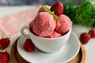 Bir fincanın içinde üç top çilekli dondurma, üzerlerinde taze bir çilek ve nane yaprağı süs olarak yerleştirilmiş. Fincan beyaz renkte ve bir tabağın üzerinde duruyor. Tabakta ve masanın üzerinde de birkaç tane taze çilek var. Arka planda hafif bulanık bir şekilde yeşil yapraklar ve pembe-beyaz bir örtü görünüyor.