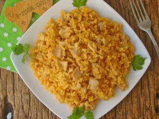 Beyaz kare bir tabakta servis edilen tavuklu bulgur pilavı. Pilavın içinde küçük küpler halinde doğranmış tavuk parçaları ve bulgur taneleri görünüyor. Pilavın rengi hafif turuncuya çalıyor, Domates veya biber salçası ile pişirilmiş. Tabakta süsleme olarak birkaç dal maydanoz var. Tabak ahşap bir masa üzerinde duruyor ve yanında metal bir çatal ile yeşil beyaz puantiyeli bir peçete yer alıyor. Peçetenin üstünde kalp şeklinde bir obje var ve üzerinde el yazısı ile bir şeyler yazıyor.