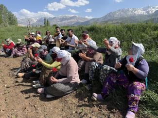 Tarlada oturan kadınlar ve birkaç erkekten oluşan bir grup. Kadınların çoğu başörtülü, şalvar giymiş ve ellerinde kırmızı karanfiller tutuyorlar. Erkekler arka sırada oturuyor, bazıları el çırpıyor. Kadınların bazıları da dua eder gibi ellerini açmış. Arka planda yemyeşil otlar, daha uzakta karlı dağlar ve mavi gökyüzü var. Hava güneşli ve açık. Grup, toprak bir yolun kenarında oturuyor. 
