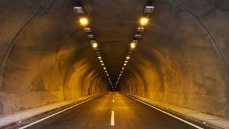 Aydınlatılmış bir karayolu tüneli. Tünelin içinden düz bir şekilde bakılıyor. Tünelin tavanında sıralı şekilde yerleştirilmiş sarı ışıklar var ve bu ışıklar tünelin içini aydınlatıyor. Yolun ortasında beyaz bir şerit bulunuyor. Yolun iki yanında kaldırım benzeri dar alanlar mevcut. Tünelin duvarları beton ve hafifçe sarımsı bir ışıkla aydınlanmış. Tünelin sonunda uzak bir noktada karanlık görünüyor, çıkış veya diğer ucu net şekilde görünmüyor. Yolda hiç araç veya insan yok, tamamen boş bir tünel.