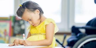 Sarı elbiseli küçük bir kız çocuğu bir masada oturuyor ve elinde bir kalemle kâğıda bir şeyler yazıp çiziyor. Saçında sarı bir toka var. Arka planda tekerlekli sandalye görünüyor. Çocuğun yüz ifadesi ciddi ve yaptığı işe odaklanmış görünüyor.