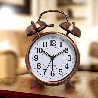 Klasik tarzda, metal çerçeveli, iki çanlı bir masa saati. Saatin kadranı beyaz ve üzerinde siyah rakamlar var. Akrep 10’u, yelkovan 2’yi, saniye ibresi 6’yı gösteriyor. Saatin üst kısmında iki adet çan ve ortasında çekiç bulunuyor. Saat, ahşap bir masa üzerinde duruyor ve arka planda bulanık bir şekilde bir koltuk ve ev ortamı görünüyor.