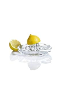 Cam limon sıkacağı ve ikiye kesilmiş bir limon. Limonun yarısı sıkacağın üstünde, diğer yarısı yanında duruyor. Arka plan tamamen beyaz ve sade. Sıkacağın camı şeffaf ve alt kısmında limon suyu birikecek şekilde tasarlanmış. Görüntüde limonun parlak sarı rengi ve camın yansımaları dikkat çekiyor.