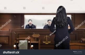 Bir mahkeme salonu. Ön planda, sırtı dönük duran uzun saçlı bir kadın var. Kadın koyu renkli bir takım elbise giymiş. Karşıda, yüksek bir kürsüde oturan iki hâkim. Hakimlerin önünde bir masa ve masanın üzerinde adalet terazisi sembolü yer alıyor. Salonun genelinde ahşap mobilyalar hâkim. Arka planda duvarda büyükçe bir pencere veya cam bölme var. Fotoğrafın sağ alt köşesinde “shutterstock” filigranı bulunuyor.