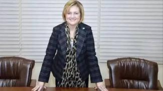 Ofis ortamında, ortada bir masa ve masanın arkasında ayakta duran İpek Atayman, iki elini masaya dayamış şekilde öne eğilmiş duruyor ve gülümsüyor. Üzerinde koyu renkli, kare desenli bir ceket ve leopar desenli bir atkı var. Arkasında iki adet kahverengi deri ofis koltuğu bulunuyor. Arka planda yatay stor perdeler kapalı görünüyor. Saçları kısa ve sarı tonlarında.