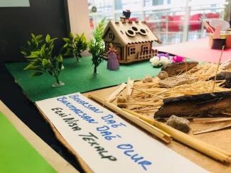 Bir maket çalışması. Sol tarafta yeşil bir zemin üzerinde iki küçük yapay ağaç ve mor elbiseli bir kadın figürü, arkada küçük, ahşap görünümlü bir ev maketi bulunuyor. Sağ tarafta samanlar, küçük taşlar ve çubuklar var. Ön planda, beyaz bir kartona mavi ve siyah kalemle yazılmış bir yazı var: "Bakarsan Bağ, Bakmazsan Dağ Olur. Ege Lisesi Teknup". Arka planda bulanık şekilde kırmızı sandalyeler ve masa görünüyor.