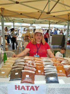 Bir pazar tezgâhı. Ön planda, üzerinde çeşitli baharatların şeffaf poşetlerde sergilendiği bir masa. Masanın arkasında, kırmızı tişört ve beyaz şapka giymiş, boynunda mavi bir yaka kartı olan Eda Nazlı duruyor. Masanın önünde "HATAY" yazılı bir afiş var. Baharat poşetleri: kırmızı, turuncu, kahverengi, siyah ve açık renkli. Masanın solunda bir yeşil cam şişe var. Arka planda başka stantlar, alışveriş yapan insanlar ve çocuklar görünüyor.