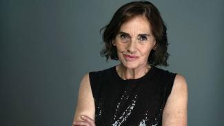 Nur Sürer. Omuz hizasında, dalgalı koyu kahverengi saçları var. Yüzünde hafif bir tebessüm ve kendinden emin bir ifade bulunuyor. Kollarını göğsünde kavuşturmuş. Siyah, parlak ve ışıltılı bir bluz giymiş. Arka plan sade, düz gri renkte. Cildi açık tenli ve yüzünde belirgin mimik çizgileri var.