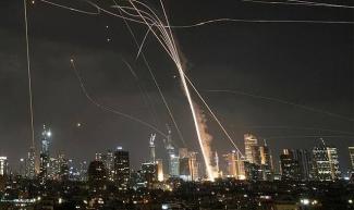 Gece vakti bir şehir silüeti ve gökyüzünde çok sayıda parlak iz bırakan roketler veya füzeler. Şehrin binaları aydınlatılmış, gökyüzünde yukarıya doğru çıkan ve bazıları eğri şekilde ilerleyen ışık izleri var. Şehrin üzerinde duman ve patlama izleri de görülüyor.