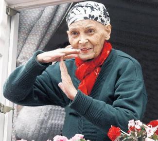 Pencere önünde duran Türkan Saylan. Başında siyah beyaz desenli bir eşarp, boynunda kırmızı bir fular, üzerinde koyu yeşil bir hırka var. Elleriyle mola anlamına gelen "T" harfi hareketi yapıyor, Yüzünde hafif bir gülümseme var. Pencerenin önünde pembe ve kırmızı çiçekler yer alıyor. Arka planda gri perdeler görünüyor.