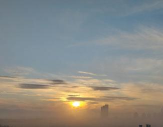 Şehir manzarası ve gün doğumu. Fotoğrafın alt kısmında sisli bir şehir silueti. Sağda yüksek bir gökdelen ve daha uzakta birkaç daha kısa bina. Gökyüzü açık mavi ve sarı tonlarında, güneş ufukta yeni doğuyor ve bulutlar hafifçe dağılmış. Güneşin ışığı sisin içinden geçerek yumuşak bir aydınlık oluşturmuş. Genel olarak sakin ve huzurlu bir sabah atmosferi var.