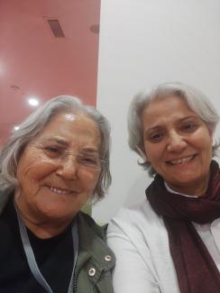 Sultan ve Nahide Hanım yan yana oturmuş, kameraya gülümseyerek selfie çekmişler. İkisinin de saçları kısa ve gri renkte. Soldaki gözlük takıyor ve koyu renk bir üstle yeşil bir ceket giymiş. Sağdaki beyaz bir üst ve bordo bir atkı takıyor. Arka planda beyaz ve pembe renkli bir duvar görünüyor. İkisi de samimi ve mutlu.