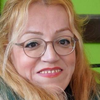 Açık tenli, sarı uzun saçlı ve gözlüklü Elif Gamze Bozo yakın plan çekimde gülümsüyor. Dudaklarında kırmızı ruj var, gözlerinde siyah eyeliner dikkat çekiyor. Arka plan parlak yeşil renkte. Yüzünde hafif bir makyaj var ve samimi bir ifadeyle kameraya bakıyor.