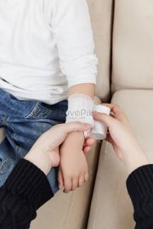 Bir çocuğun koluna bandaj sarılıyor. Çocuk beyaz bir uzun kollu tişört ve mavi kot pantolon giymiş. Bir yetişkin, çocuğun bileğine veya ön koluna dikkatlice beyaz bir bandaj sarıyor. İkisi de bir koltukta oturuyor.