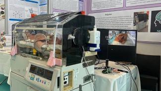 Bir kuvöz ve yanında bir bilgisayar monitörü. Kuvözün içinde bir bebek yatıyor. Kuvözün dışına çeşitli tıbbi cihazlar ve sensörler yerleştirilmiş. Monitörde kuvözün içindeki bebeğin görüntüsü yer alıyor. Arka planda ise bilimsel bir poster ve yazılar var.