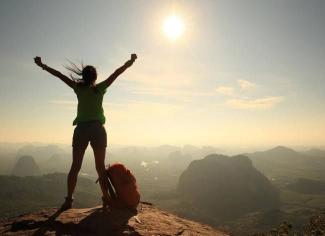 Dağın zirvesinde duran bir kadın silueti, kollarını zafer işaretiyle yukarı kaldırmış. Kadının yanında turuncu bir sırt çantası var. Güneş gökyüzünde parlarken, arka planda sisli dağlar ve geniş bir vadi manzarası. Kadının saçları rüzgârda savrulmuş.