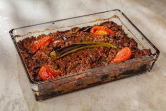 Fırın tepsisinde hazırlanmış bir yemek. Üst kısmında kıyma harcı, dilimlenmiş domatesler ve bütün bir yeşil biber yer alıyor. Alt katmanda kızartılmış patlıcan dilimleri görünüyor. Yemeğin kenarları hafifçe kızarmış. Cam fırın kabında servis edilmiş ve oldukça iştah açıcı görünüyor.