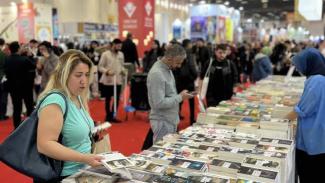 Kitap fuarında kalabalık bir ortam. Ön planda, masaların üzerinde üst üste dizilmiş birçok kitap var. Sol tarafta açık mavi tişörtlü bir kadın kitaplara bakıyor ve bir kitabı eline almış. Ortada gri gömlekli bir adam telefonuna bakıyor. Sağda mavi başörtülü bir kadın kitaplara göz atıyor. Arka planda fuar alanında dolaşan ve kitaplara bakan birçok insan var. Zemin kırmızı halı kaplı.