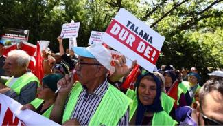 Protesto yapan bir grup insan. Çoğu kişi fosforlu yeşil yelek giymiş. Ellerinde Türk bayrakları ve çeşitli pankartlar var. En önde bir adam "TALANA DUR DE!" yazılı bir pankart tutuyor. Arka planda ağaçlar ve güneşli bir hava. İnsanlar kararlı ve ciddi. Pankartlarda çevreyle ve doğayla ilgili mesajlar yer alıyor.