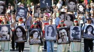 Bir grup insanın açık havada yürüyüş yaptığı bir fotoğraf. Ön safta yer alanlar, ellerinde büyük boyutlu siyah-beyaz portre fotoğrafları taşıyor. Fotoğrafların altında "ONURUMUZ" yazısı ve kişilerin isimleri yer alıyor. Yürüyüşe katılan bazı kişiler başlarına kırmızı ve yeşil bantlar takmış. Arkada kalabalık bir grup daha var.