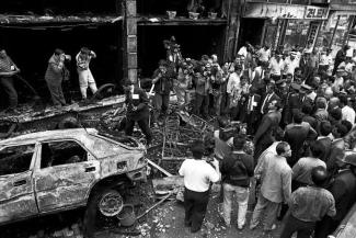 Yanmış bir araba, enkaz ve hasar görmüş bir bina önünde toplanmış kalabalık bir insan grubu. Olay yerinde çok sayıda gazeteci, polis ve meraklı vatandaş var. Fotoğraf siyah-beyaz. İnsanlar olay yerini inceliyor, bazıları fotoğraf çekiyor. Arka planda dükkân tabelaları ve yanmış bir vitrin.