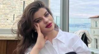 Beyaz gömlek giymiş, uzun koyu saçlı Ayşe Tokyaz, başını eline yaslamış şekilde poz veriyor. Arka planda camdan dışarıya bakıldığında deniz ve bazı binalar görünüyor. Mekân ferah ve aydınlık, arka planda taş duvar ve açık hava detayları mevcut. Tokyaz’ın makyajı belirgin, dudakları koyu kırmızı renkte. Genel olarak rahat ve düşünceli bir pozda.