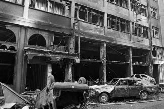 Yıkılmış bir bina ve hasar görmüş arabalar. Görüntü siyah beyaz. Binanın alt katı tamamen tahrip olmuş, camlar kırılmış ve üst katlarda da ciddi hasar var. Ön planda bir araba ters dönmüş, diğer arabalar ezilmiş ve yanmış gibi görünüyor. Etrafta enkazlar ve döküntüler bulunuyor.