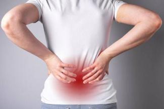 Sırt ağrısı hisseden bir kişi beyaz bir tişört ve açık renk pantolon giymiş. İki elini belinin alt kısmına koymuş ve bel bölgesinin tam ortasında kırmızı bir renk efekti var, bu bölgede ağrı veya rahatsızlık olduğunu gösteriyor. Kolları iki yana açılmış şekilde duruyor ve kişi arkadan görülüyor. Arka planda düz, açık gri bir duvar var.