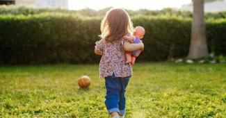 Bahçede yürüyen küçük bir çocuk. Saçları açık ve üstünde çiçek desenli bir bluz ile mavi bir pantolon var. Kucağında bir oyuncak bebek tutuyor. Çocuk çimenlerin üzerinde, arka planda toprağa dikilmiş bir ağaç ve koyu yeşil çalılar görülüyor. Yerde bir tane turuncu renkli top var. Güneş ışığı arkadan geliyor ve ortam aydınlık görünüyor. Çocuk, kameraya sırtı dönük şekilde yürüyor.