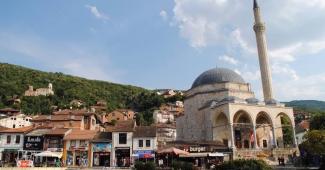 Tarihi bir cami ve çevresindeki dükkanlar. Görüntünün ortasında büyük kubbeli ve minareli bir cami. Caminin önünde üç kemerli bir giriş bulunuyor. Caminin yanında ve önünde çeşitli dükkanlar sıralanmış; bazı dükkanlarda tabelalar ve şemsiyeler görülüyor. Cami, klasik Osmanlı mimarisi tarzında yapılmış. Arka planda ormanlık bir tepe ve tepenin yamaçlarına dağılmış evler var. Gökyüzü açık ve bulutlu.