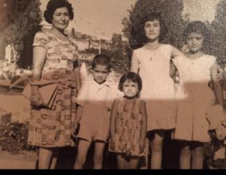 Siyah beyaz, eski bir aile fotoğrafı. Solda yetişkin bir kadın, yanında dört çocuk. Kadının elinde bir dosya ya da evrak var. Çocuklardan biri erkek (şortlu ve gömlekli), diğer üçü kız çocuğu (elbiseli). Hepsi yan yana durmuş ve kameraya bakıyor. Arka planda ağaçlar ve biraz net olmayan şehir manzarası var.