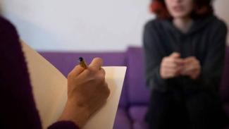 Psikolojik danışmanlık ortamı. Ön planda bir kişi elinde kalemle kâğıda bir şeyler yazıyor. Arka planda karşısında oturan ve ellerini birleştirmiş genç bir kadın bulanık şekilde görülüyor. Kadın koyu renk bir üst giymiş, mor koltukta oturuyor.