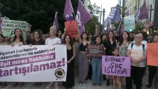 Kadınların düzenlediği bir protesto yürüyüşü. Kalabalık grup yaya bir caddede toplanmış ve ellerinde çeşitli pankartlar ve mor bayraklar taşıyorlar. Pankartlarda, "Diyanet hutbeleriyle kazanımlarımıza saldırıyor! Eşit miras hakkımız tartışılamaz!", "Diyanet suç işliyor", "Diyanet kadınların kazanımlarına saldırarak suç işliyor", "Haklarımızdan ve kazanımlarımızdan vazgeçmiyoruz." Yazısı. Kadınlar ciddi bir ifadeyle kameraya bakıyor. Arka planda mor kadın hakları bayrakları dikkat çekiyor.