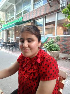 Dış mekânda bir kafe ya da restoran önünde oturanNazalin Selvi. Üzerinde kırmızı renkte, siyah puantiyeli ve fırfırlı bir bluz var. Kulağında yıldız şeklinde sallantılı bir küpe takıyor. Saçları toplanmış ve yüzünde hafif bir tebessüm var. Arka planda kafenin dış kısmı, sandalyeler, masalar ve saksı bitkileri görülüyor.