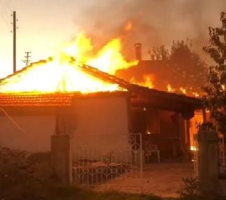 Alevler içinde bir ev. Çatı kısmından yoğun alevler ve dumanlar yükseliyor. Yangın çok şiddetli ve üst kat tamamen yanıyor. Evin önünde küçük bir bahçe ve beyaz bir kapı var. Arka planda ağaçlar seçiliyor. Yangın alanında görünür şekilde kimse yok.