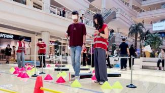 Bir alışveriş merkezinin zemin katında kurulan bir parkur. Yerde renkli koniler ve engeller yer alıyor. Bir adamın gözleri bez bir bantla kapalı ve bir kadın kolundan tutup ona rehberlik ediyor; kadın kırmızı-beyaz çizgili bir yelek giymiş. Arkada başka insanlar da benzer şekilde parkurda ilerliyor ve bazıları yine elinde baston tutuyor. Çevrede alışveriş yapan insanlar, dükkanlar ve oturanlar var. Mekân aydınlık ve ferah görünüyor.