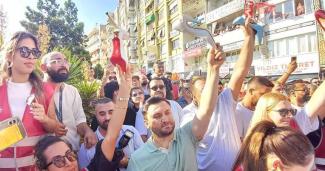 Kalabalık bir grup insan açık havada, bir pek çoğu ellerinde tek bir yüksek topuklu ayakkabı tutuyor. İnsanlar neşeli ve kararlı, bazıları güneş gözlüğü takıyor. Arka planda çok katlı apartmanlar ve dükkân tabelaları. Güneş parlıyor.