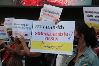 Önde, elinde pankart tutan bir kadın, siyah maske takıyor ve kot ceket giymiş. Tuttuğu pankartta büyük harflerle "FETVALAR SİZİN SOKAKLAR BİZİM OLSUN!" yazıyor. Alt kısmında “ekmek ve gül” ifadesi ile bir çiçek görseli. Arka planda başka katılımcılar ve farklı pankartlar görülüyor. Diğer pankartlarda "Diyanete Değil Kadınlara Bütçe!", "Kiralar Denetlensin Zamlar Geri Alınsın!" ve "Parasız Barınma Güvenli Yurt İstiyoruz" gibi çeşitli talepler yer alıyor.