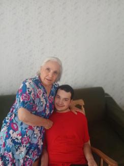 Menekşe Koçak ve oğlu. Koçak, çiçek desenli mavi ve pembe renkli bir elbise giymiş ve gri kısa saçlı. Yanında duran oğlu Koray, kırmızı bir tişört giymiş ve bir sandalyede oturuyor. Koçak elini sevgiyle oğlunun omzuna koymuş. Arkada açık renkli desenli bir duvar kâğıdı ve koyu yeşil bir kanepe bulunuyor. İkisi de kameraya bakıyor ve hafifçe gülümsüyor.