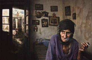 Yaşlı bir kadın, eski ve yıpranmış bir odada, başında siyah bir örtüyle oturuyor. Elinde sigara var ve yüzü derin kırışıklıklarla dolu, oldukça yorgun ve düşünceli görünüyor. Üzerinde mor bir hırka var. Odanın duvarları solmuş ve kirli, duvarda eski fotoğraflar ve çerçeveler asılı. Kadının oturduğu kanepe veya divan üzerinde dağınık bir örtü var. Arka planda açık bir kapıdan dışarı bakılıyor; dışarıda bitkiler ve eski, taş bir duvar, bir de keçi veya koyun görünüyor. Gökyüzünde uçan kuşlar var.