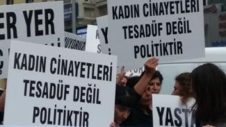 Bir grup insan ellerinde pankartlarla bir protesto veya yürüyüşte bulunuyorlar. Pankartların üzerinde büyük harflerle yazılmış yazılar bulunuyor. En öndeki pankartlarda, “KADIN CİNAYETLERİ TESADÜF DEĞİL POLİTİKTİR”. Yazıyor.