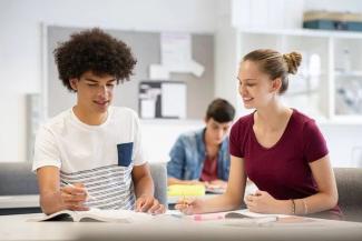 Sınıf ortamında birlikte ders çalışan iki genç. Ön planda, biri kısa kollu beyaz tişört giymiş kıvırcık saçlı bir erkek ve diğeri bordo tişört giymiş sarışın bir kız oturuyor. İkisi de masada açık kitaplara ve defterlere bakıyorlar, aralarında bir etkileşim ve anlaşma var; kız öğrencinin elinde kalem var ve gülümsüyor. Arka planda başka bir öğrenci ders çalışıyor.