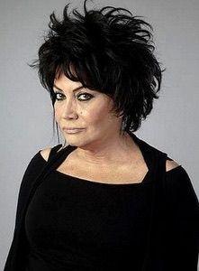 Anta Toros. Saçları siyah ve kabarık, dağınık bir şekilde şekillendirilmiş. Yüzünde makyaj var; özellikle göz makyajı belirgin ve göz çizgileri vurgulanmış. Üstünde siyah bir kıyafet var; omuz kısmında açıklıklar bulunuyor. Hafif yana dönük ve ciddi, kendinden emin bir ifadeyle bakıyor. Arka plan düz ve gri renkte.