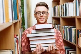 Kütüphane rafları arasında duran, gözlük takan genç bir erkek. Üzerinde kırmızı-beyaz ekose bir gömlek var ve kucağında altı tane kitap tutuyor. Gülümsüyor ve doğrudan kameraya bakıyor. Arka planda iki yanında kitap dolu raflar görünüyor.
