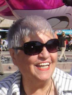 Ayten Gündüz’ün açık havada çekilmiş, gülen portresi. Kısa, gri saçlı, büyük siyah güneş gözlüğü takıyor ve kırmızı ruj sürmüş. Üzerinde beyaz zemin üzerine ince çizgili, açık renkli bir gömlek var. Arkada, park veya meydanda insanlar ve yerde güvercinler görünüyor. Üst kısımda pembe ve büyük bir balon veya oyuncak objenin bir kısmı yer alıyor.