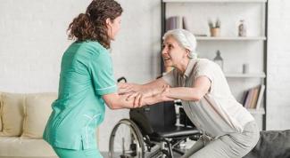 Oturmaya çalışan yaşlı bir kadın ile ona yardım eden sağlık personeli. Kadın beyaz saçlı ve açık renk kıyafetler giymiş, sağlık personeli yeşil üniforma içinde. Arkalarında tekerlekli sandalye ve kitaplık bulunuyor. Arka planda bir koltuk var. Sağlık personeli yaşlı kadının ellerini tutarak ona destek oluyor.