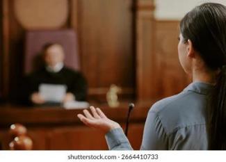 Mahkeme salonunda bir kadın, hâkimin önünde konuşuyor. Kürsünün önünde duruyor ve elini hafifçe yukarı kaldırmış. Arka planda bir hâkim, mahkeme cübbesiyle oturuyor ve elinde bazı belgeler tutuyor. Masanın üstünde küçük bir adalet heykelciği var.