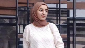 Açık renkli, kareli desenli bir kazak giymiş ve başında açık kahverengi bir başörtüsü olan Munise Demirer. Kafesli metal bir yapının önünde ayakta duruyor ve sola doğru bakıyor. Arka planda cam pencereler ve kahverengi- siyah çerçeveler görünüyor. Demirer’in yüzünde sakin bir ifade var.