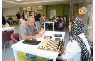 Bir masa etrafında satranç oynayan iki kişi. Erkek oyuncu hamle yapmaya hazırlanıyor, kadın oyuncu Fariba’nın başında siyah beyaz puantiyeli bir başörtüsü var ve dikkatle oyunu izliyor. Masada satranç tahtası, satranç taşları dizili ve bir satranç saati bulunuyor. Arka planda başka masalarda da insanlar oturmuş, bazıları satranç oynuyor, bazıları sohbet ediyor. Oda ferah, modern ve renkli sandalyeler dikkat çekiyor. Arka duvarda "Özgün" yazılı bir afiş ve yanında su sebili var. 